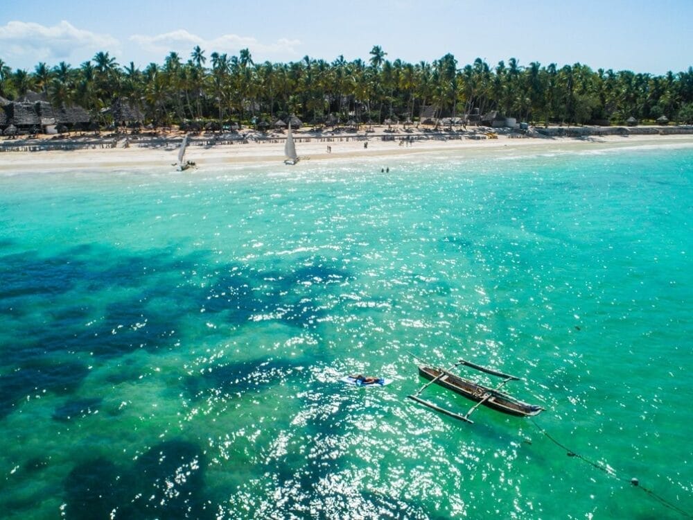 Una piccola barca di legno galleggia sull'acqua limpida e turchese vicino a una spiaggia sabbiosa di Zanzibar fiancheggiata da palme, con alcune persone e capanne del villaggio di Zanzibar visibili lungo la riva sotto un cielo luminoso.