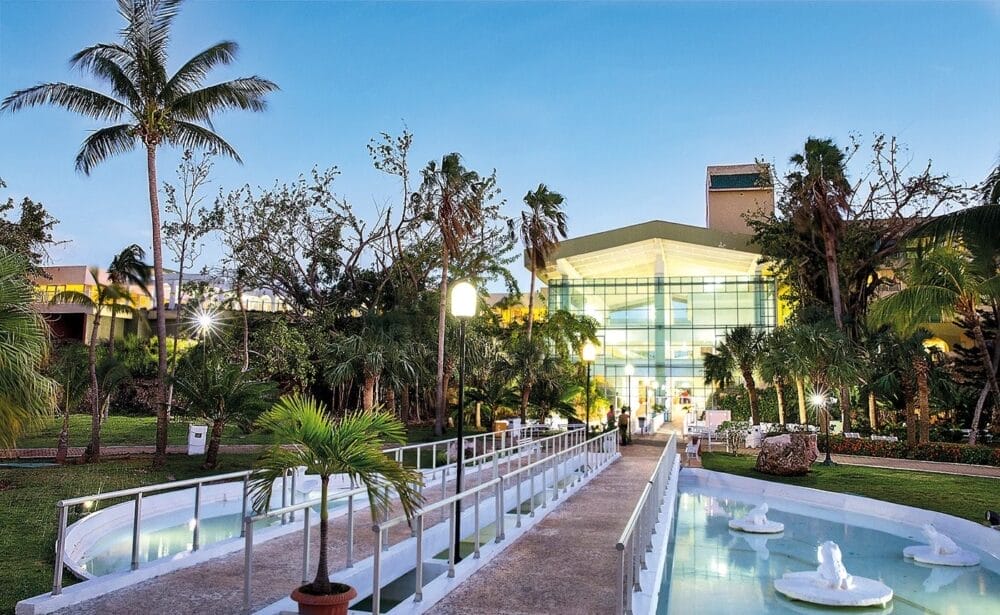 Una passerella con ringhiere bianche attraversa un gioco d'acqua decorativo e conduce al moderno Veraclub Starfish Varadero, circondato da palme e da una vegetazione lussureggiante: uno scenario invitante per memorabili vacanze a Cuba sotto un cielo azzurro e limpido al tramonto.