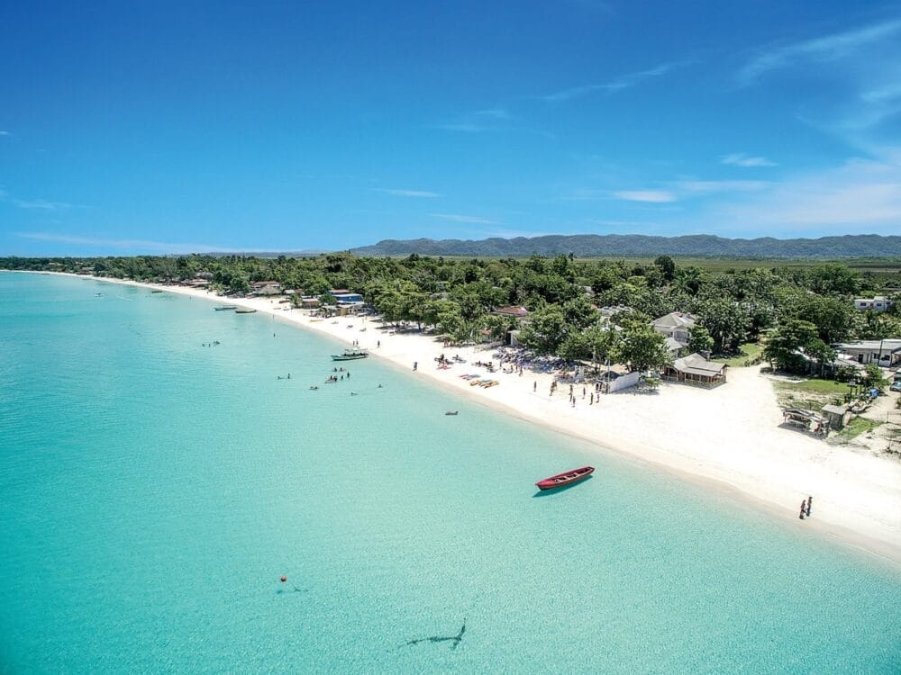 Veduta aerea del resort Veraclub Negril in Giamaica: acqua limpida e turchese, sabbia bianca, persone che nuotano e si rilassano, una barca rossa nell'acqua, alberi verdeggianti e montagne lontane sotto un cielo blu brillante.
