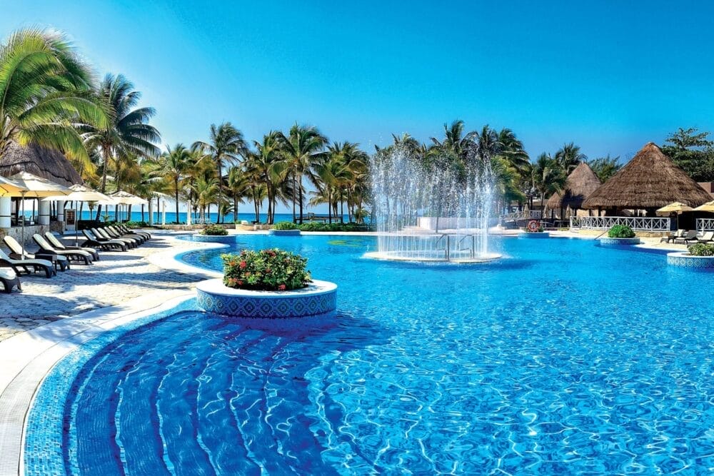 La grande piscina blu scintillante del Veraclub Royal Tulum è caratterizzata da una fontana centrale, circondata da sedie a sdraio, ombrelloni, palme e cabine con tetto di paglia sotto un cielo limpido in questo hotel all-inclusive vicino all'oceano.