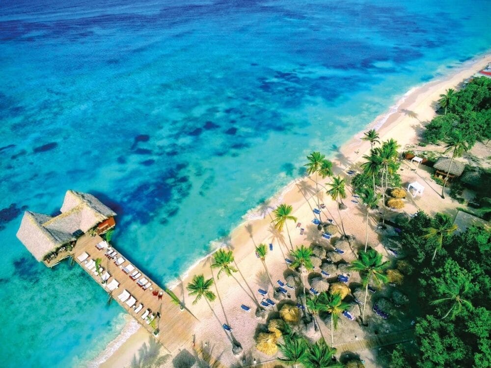 Vista aerea di una spiaggia tropicale del Sunscape Dominicus, con acqua turchese, palme, arenile sabbioso e un molo di legno che conduce a capanne con tetto di paglia. Sedie a sdraio e ombrelloni sono sparsi sul lungomare.