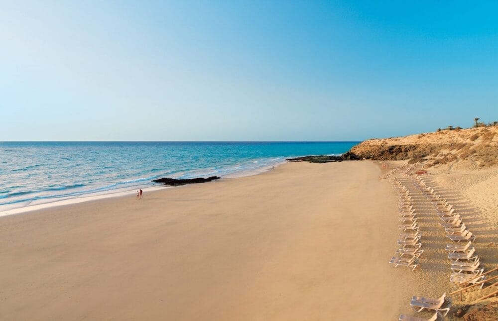 Ampia spiaggia sabbiosa a Fuerteventura con file di sedie a sdraio vuote sulla destra, mare azzurro e calmo sulla sinistra e alcune persone che passeggiano vicino alla riva sotto un cielo limpido. Affioramenti rocciosi incorniciano lo sfondo vicino al Veraclub Tindaya.