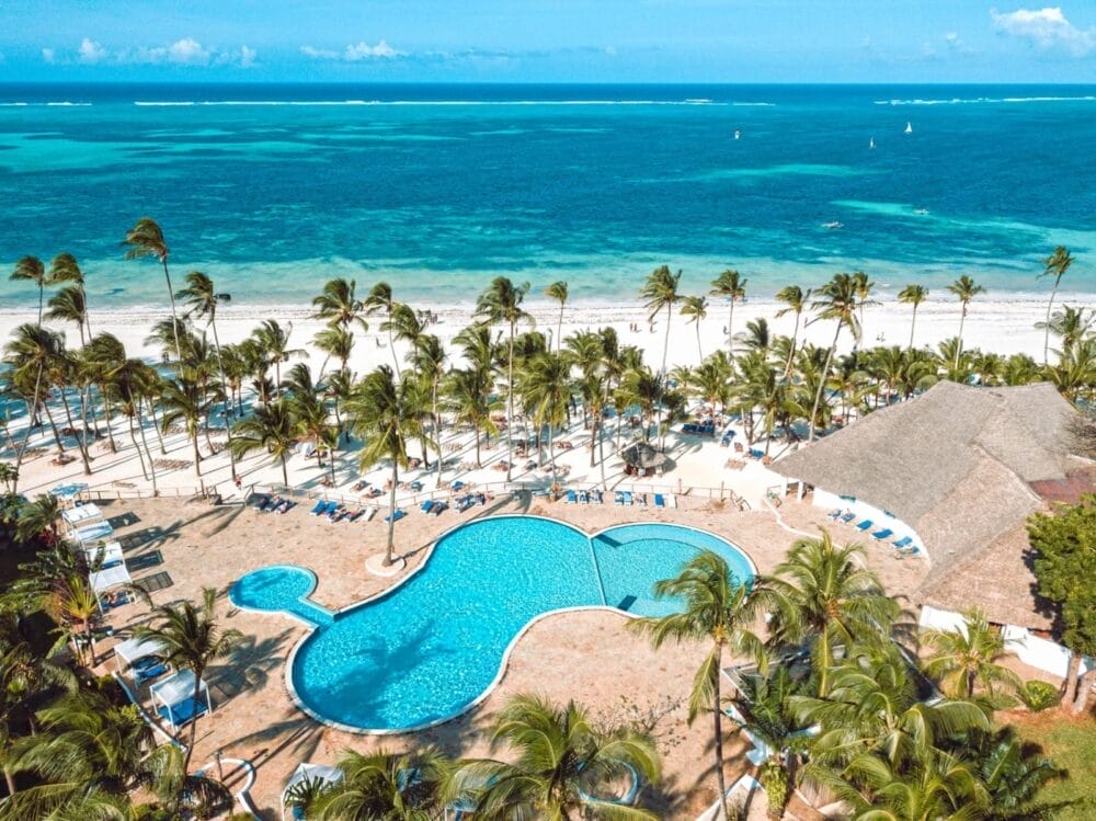 Vista aerea del Bravo Kiwengwa, un resort sulla spiaggia tropicale con una grande piscina dalla forma unica, circondata da palme, sedie a sdraio e sabbia bianca, che si affaccia sull'oceano turchese e sul cielo azzurro.
