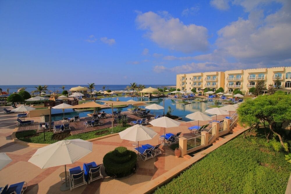 Un resort con una piscina circondata da sedie a sdraio e ombrelloni bianchi, un paesaggio verde e palme. Sullo sfondo, il Veraclub Salalah offre una spiaggia sabbiosa, l'oceano e un edificio dell'hotel sotto un cielo blu con nuvole sparse.