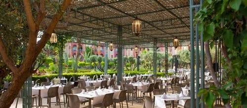 Patio del ristorante all'aperto dell'Iberostar Palmeraie Marrakech, con tavoli disposti sotto un pergolato ornato di lanterne. Il verde lussureggiante e gli alberi creano un'atmosfera serena e invitante, perfetta per cenare negli splendidi dintorni di Marrakech.