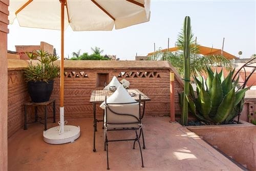 Un piccolo patio all'aperto dell'Angsana Riads di Marrakech presenta un tavolo e due sedie sotto un grande ombrellone bianco, circondato da piante in vaso, un cactus, pareti decorative in pietra e un cielo limpido sullo sfondo.
