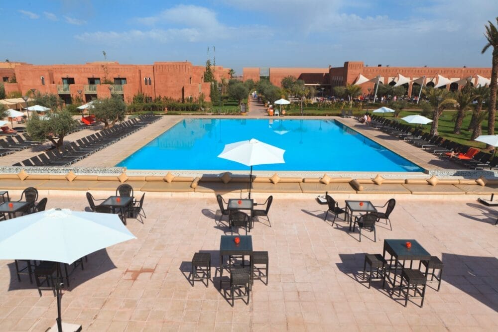 Grande piscina all'aperto del Kenzi Agdal, circondata da sedie a sdraio, tavoli ombreggiati e ombrelloni, con edifici color terracotta e prati verdi sullo sfondo sotto il cielo parzialmente nuvoloso della Medina.