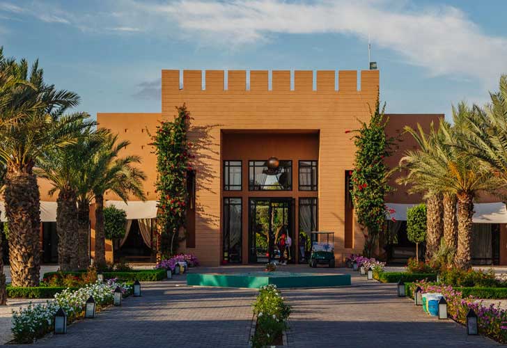 L'Aqua Mirage Marrakech è un edificio moderno con un tetto piatto e merlato, circondato da palme e fiori colorati; una piccola fontana circolare abbellisce il cortile che conduce all'ingresso.