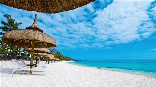 All'Ambre Sun Resort vi attende una spiaggia tropicale con sabbia bianca, acqua turchese e ombrelloni di paglia che ombreggiano sedie a sdraio bianche, il tutto incorniciato da palme sotto un cielo azzurro parzialmente nuvoloso.
