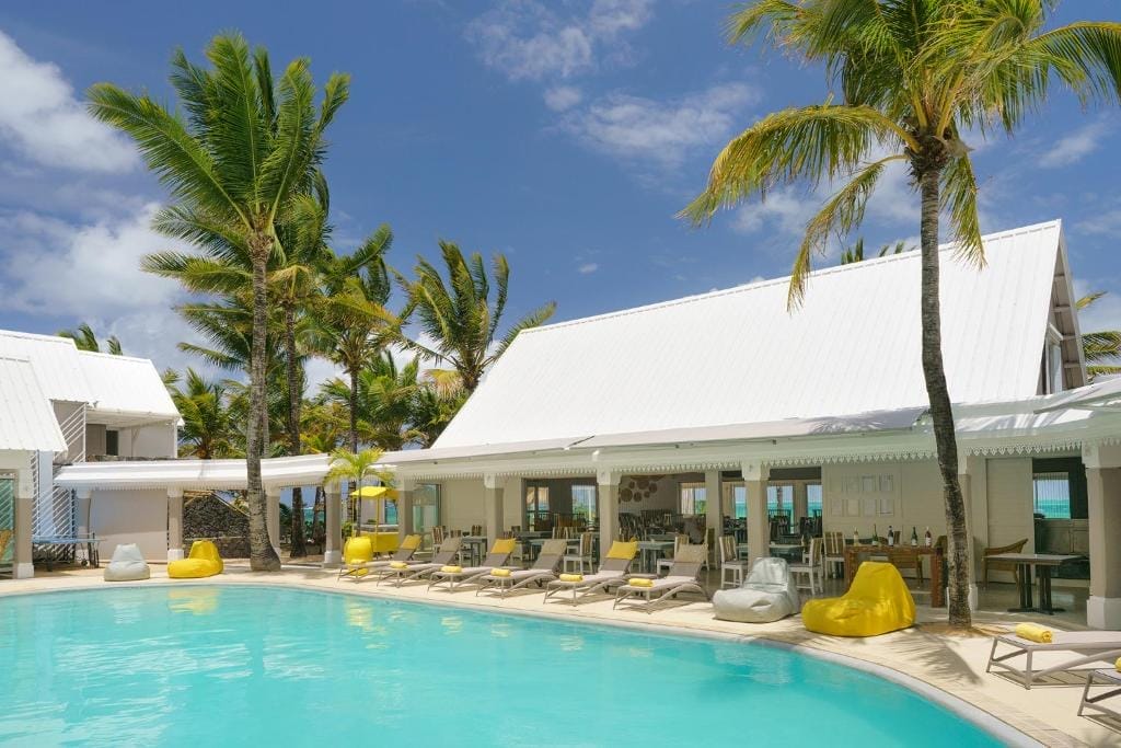 Il Tropical Attitude è un resort tropicale di Mauritius con una piscina circondata da palme, sedie a sdraio gialle e bianche, sacchi di fagioli ed edifici bianchi con tetti spioventi sotto un cielo blu brillante, perfetto per una fuga per soli adulti.
