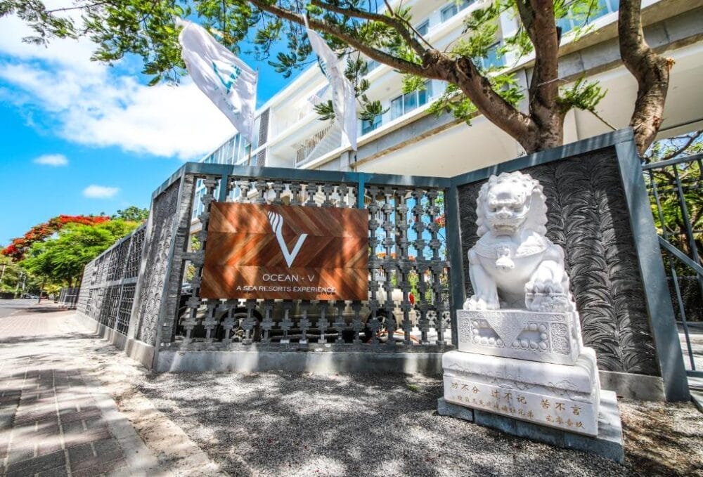 Ingresso dell'Ocean V Hotel, un resort, caratterizzato da un cancello decorativo, un'insegna con il nome dell'hotel, una statua di leone bianco e bandiere, circondato da alberi sotto un cielo azzurro e soleggiato.