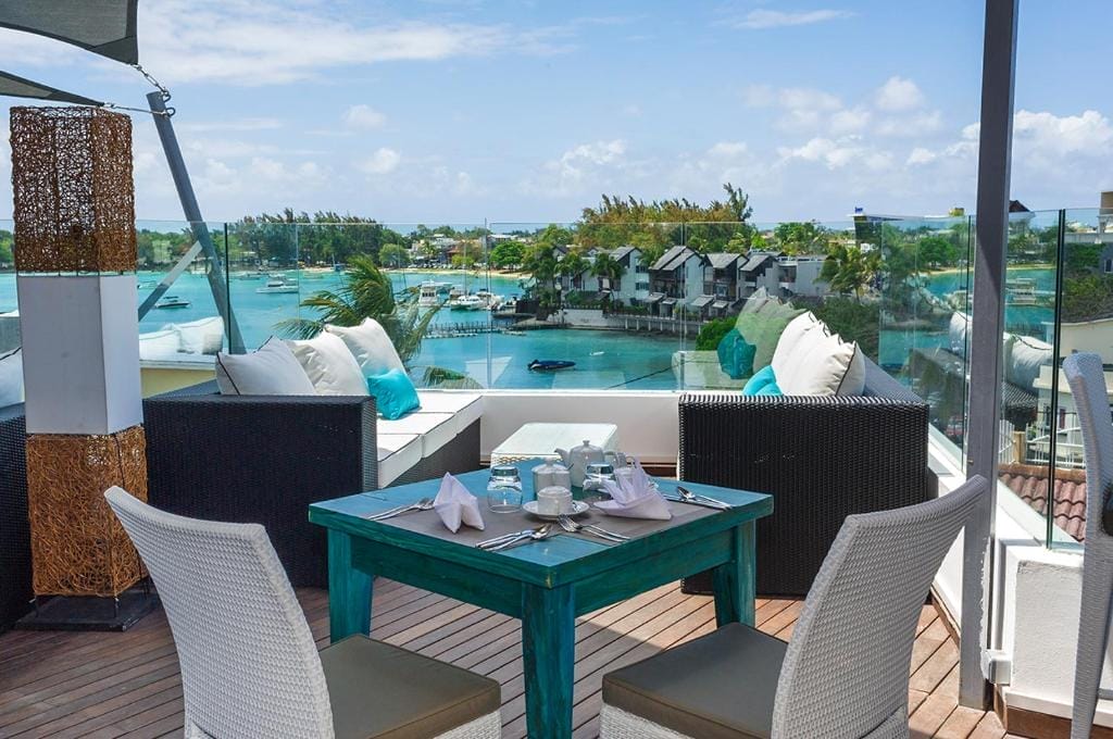 La zona pranzo sul tetto dell'Azur Paradise è caratterizzata da un tavolo turchese e sedie bianche, con vista su un porticciolo con barche, ville sul lungomare e acqua cristallina sotto un cielo soleggiato. Sedute con cuscini e ringhiere in vetro migliorano l'atmosfera rilassante.