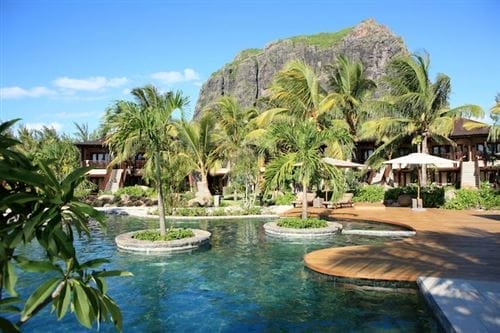 La piscina del resort tropicale Lux Le Morne è circondata da palme, terrazze in legno e vegetazione lussureggiante, con una montagna ripida e rocciosa che si erge sullo sfondo sotto un cielo blu con nuvole sparse.