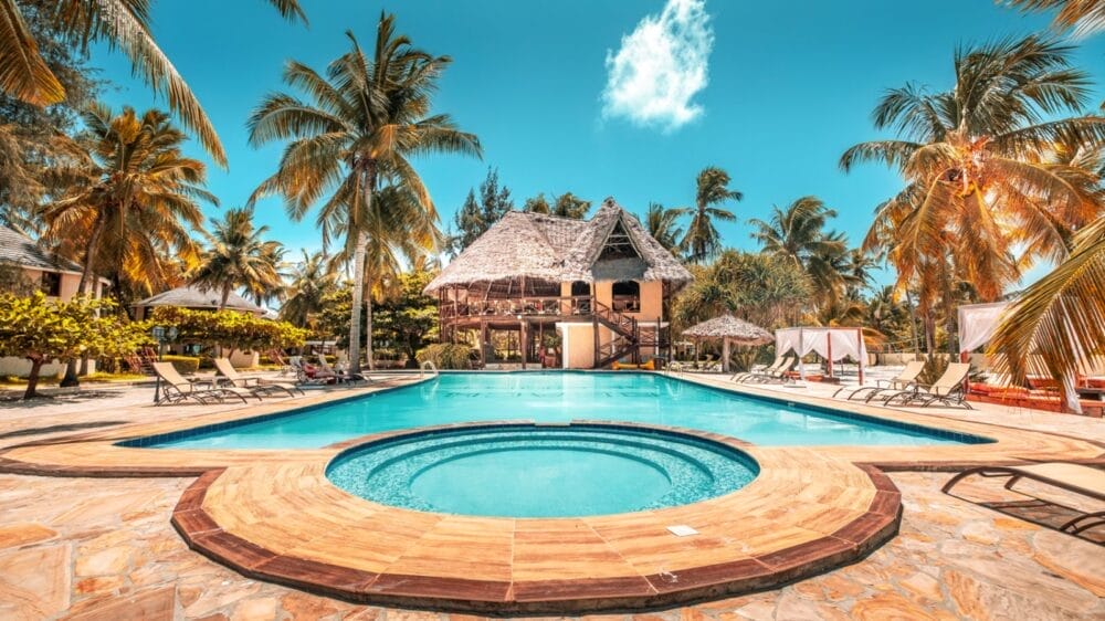 Al Club Coralia Kae Beach Zanzibar, un resort tropicale dispone di una vasca idromassaggio circolare e di un'ampia piscina circondata da palme, lettini ed edifici con tetti di paglia sotto un cielo blu brillante.