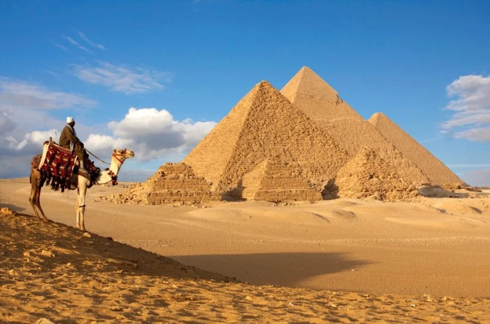 Una persona in sella a un cammello addobbato con coperte colorate guarda le tre Piramidi di Giza sotto un cielo azzurro, circondato dalla sabbia del deserto: l'inizio perfetto di una Crociera Nefertari prima di esplorare Il Cairo.