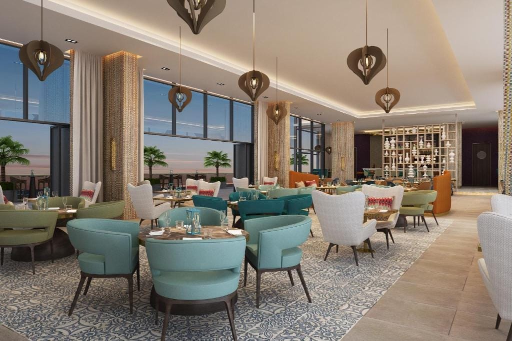 Un ristorante moderno ed elegante del Centara Mirage Beach Resort, con poltrone color verde acqua e beige, tavoli rotondi per mangiare, grandi finestre con palme e tramonto, pavimenti a motivi e eleganti luci a sospensione appese al soffitto.