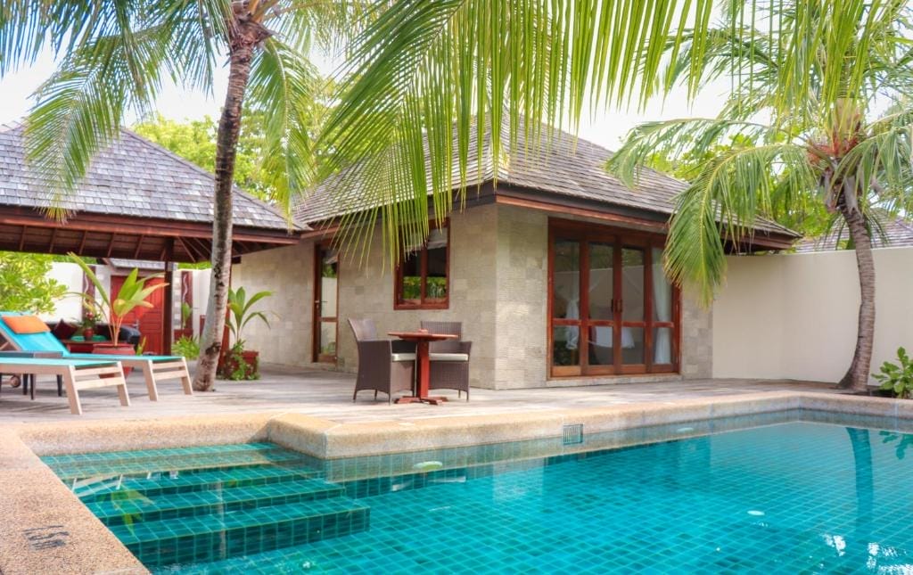 Una villa tropicale con piscina privata nel resort di lusso di Kuredu Island Resort, alle Maldive. Circondata da palme, presenta porte in legno, un tetto di paglia, sedie a sdraio e un piccolo tavolo nel patio per una perfetta fuga dall'isola.