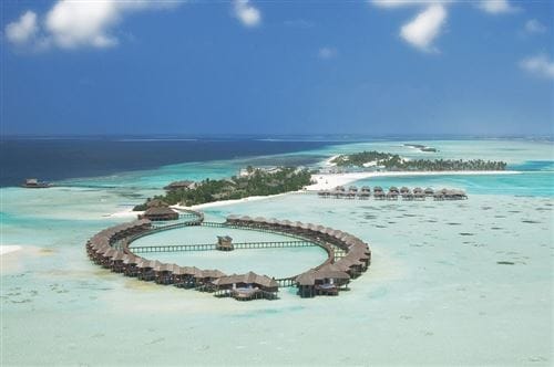 Vista aerea di un resort overwater con ville disposte circolarmente sopra acque turchesi e trasparenti, circondate da spiagge di sabbia bianca e da una vegetazione lussureggiante sotto un cielo blu brillante: un'idilliaca fuga automatica di Bozza.