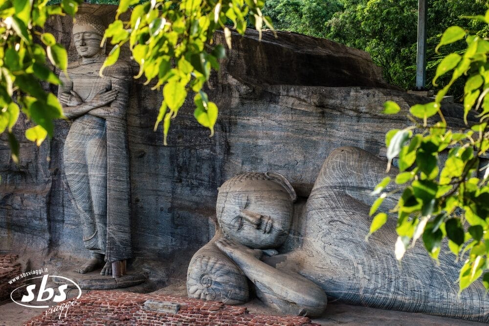 Grandi statue in pietra di un Buddha sdraiato e di una figura in piedi, parzialmente incorniciate da foglie verdi, poggiano su uno sfondo roccioso in un antico sito. L'immagine include il logo "sbs" nell'angolo inferiore sinistro ed è contrassegnata come Bozza automatica.