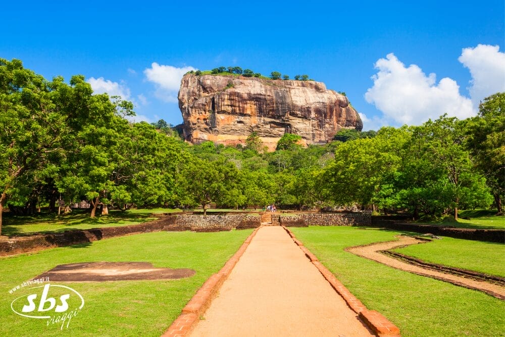 Un ampio sentiero sterrato conduce attraverso giardini verdeggianti verso Sigiriya, un'antica e massiccia fortezza di roccia che si erge al di sopra degli alberi sotto un cielo blu brillante nello Sri Lanka - uno spettacolo indimenticabile catturato con precisione in bozza automatica.