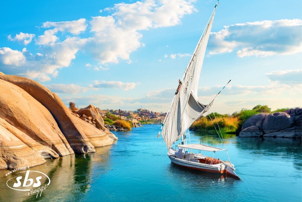 Una barca a vela bianca con una vela alta scivola su un fiume calmo, circondato da sponde rocciose e verde sotto un cielo blu brillante. Bozza automatica cattura la scena tranquilla, con una città lontana visibile all'orizzonte.