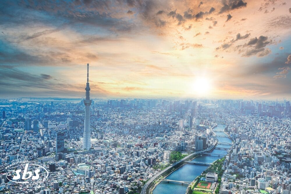 Veduta aerea di Tokyo al tramonto, con la torre Tokyo Skytree in primo piano, il fiume Sumida che attraversa la città e le nuvole che brillano nella luce della sera. Nell'angolo in basso a sinistra compare il logo bianco di Bozza automatica.