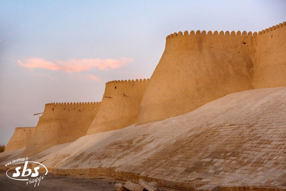 Mura storiche di una fortezza in pietra beige con merli, fotografate al tramonto sotto un cielo pastello. Il muro si incurva in primo piano. Il logo di Bozza automatica e la scritta "sbs insight" compaiono nell'angolo in basso a sinistra.