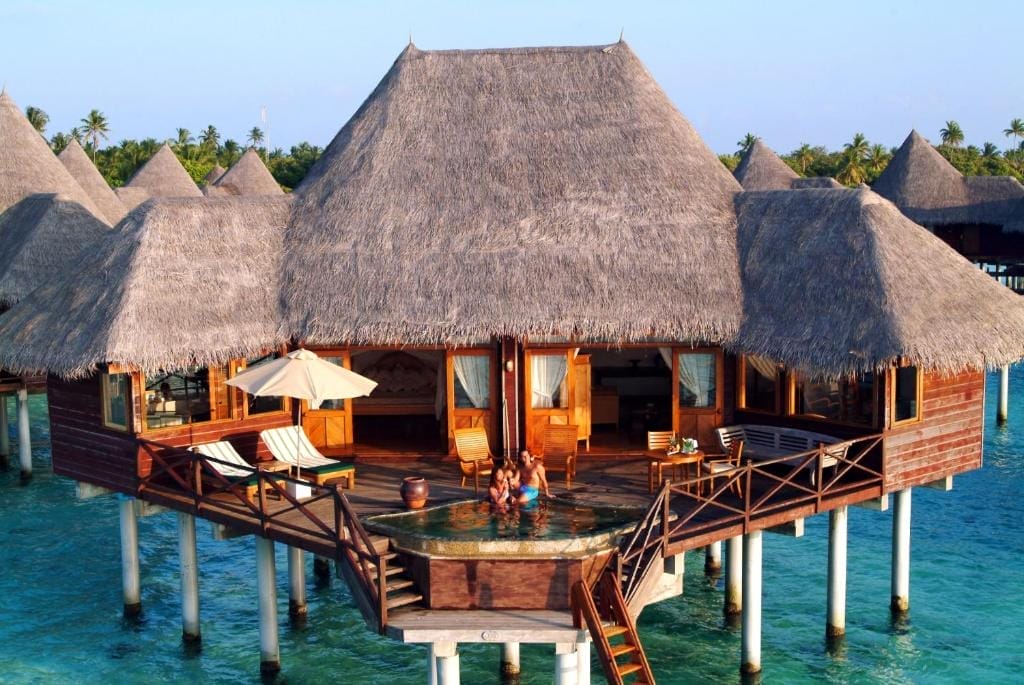 Un bungalow overwater con tetto in paglia del resort Coco Palm Dhuni Kolhu Maldives si trova sopra l'acqua turchese. Sul ponte si trovano sedie a sdraio e un ombrellone, mentre gli ospiti si rilassano in una piscina privata con vista sull'oceano.