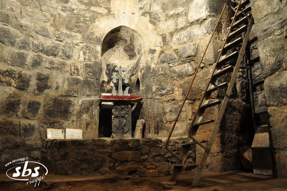 Una camera millenaria con pareti in pietra e un piccolo altare con una croce di metallo, targhe e una scala appoggiata al muro. L'ambiente evoca il passato storico dell'Armenia, con pavimenti in pietra grezza e luci soffuse.