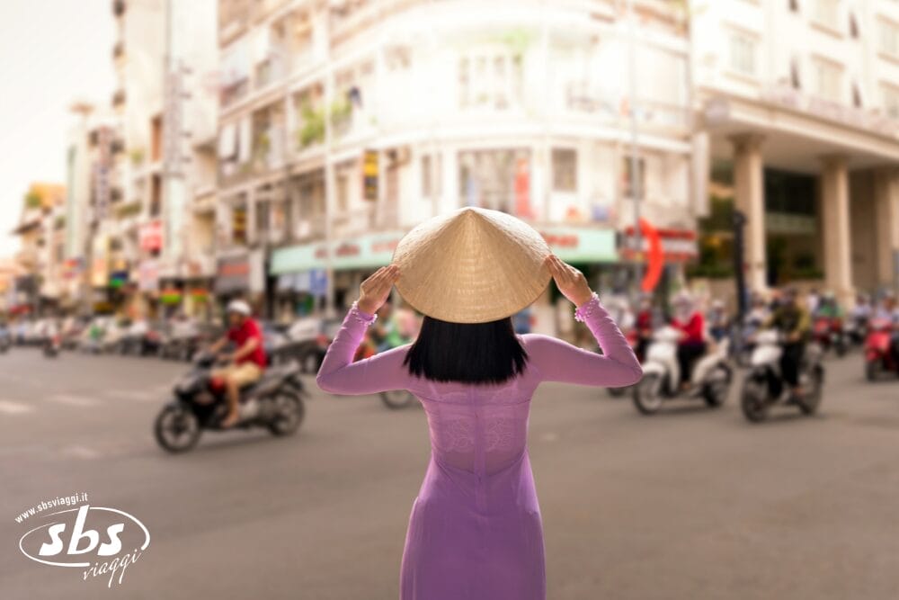 Una donna con un abito viola e un cappello conico si trova di spalle all'obiettivo, osservando una strada trafficata piena di motociclette e di edifici sfocati: una vera e propria bozza automatica della vita urbana.