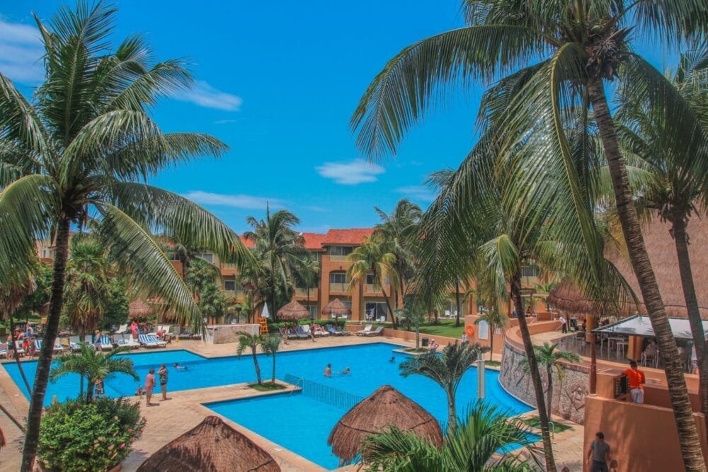 Un resort tropicale, il Bravo Viva Azteca, dispone di una grande piscina all'aperto circondata da palme, sedie a sdraio e ombrelloni di paglia. Diverse persone si rilassano a bordo piscina con edifici colorati sullo sfondo, sotto un cielo blu brillante.