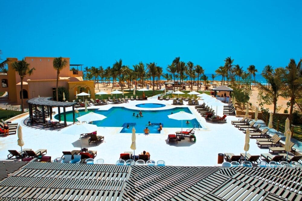 Un'area della piscina del Salalah Resort con persone che nuotano e si sdraiano, circondate da palme e lettini, con la spiaggia e l'oceano visibili sotto un cielo blu chiaro sullo sfondo.