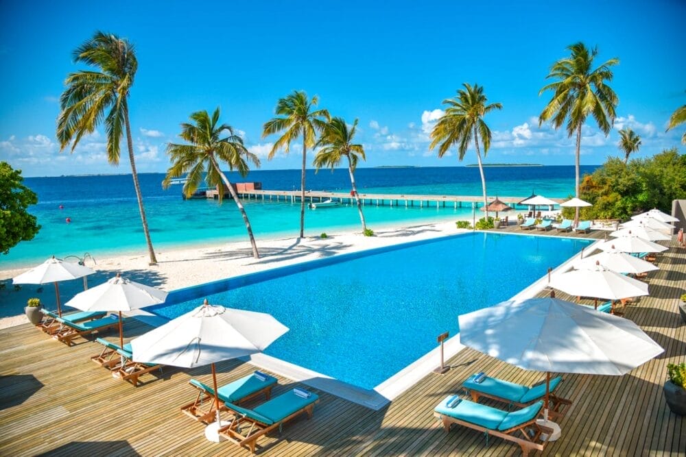 Resort di lusso alle Maldive, il Reethi Faru Resort vanta una piscina a sfioro, lettini sotto ombrelloni bianchi, alte palme e una spiaggia sabbiosa che conduce all'oceano blu, mentre un molo di legno si protende nell'acqua sotto un cielo luminoso e soleggiato.
