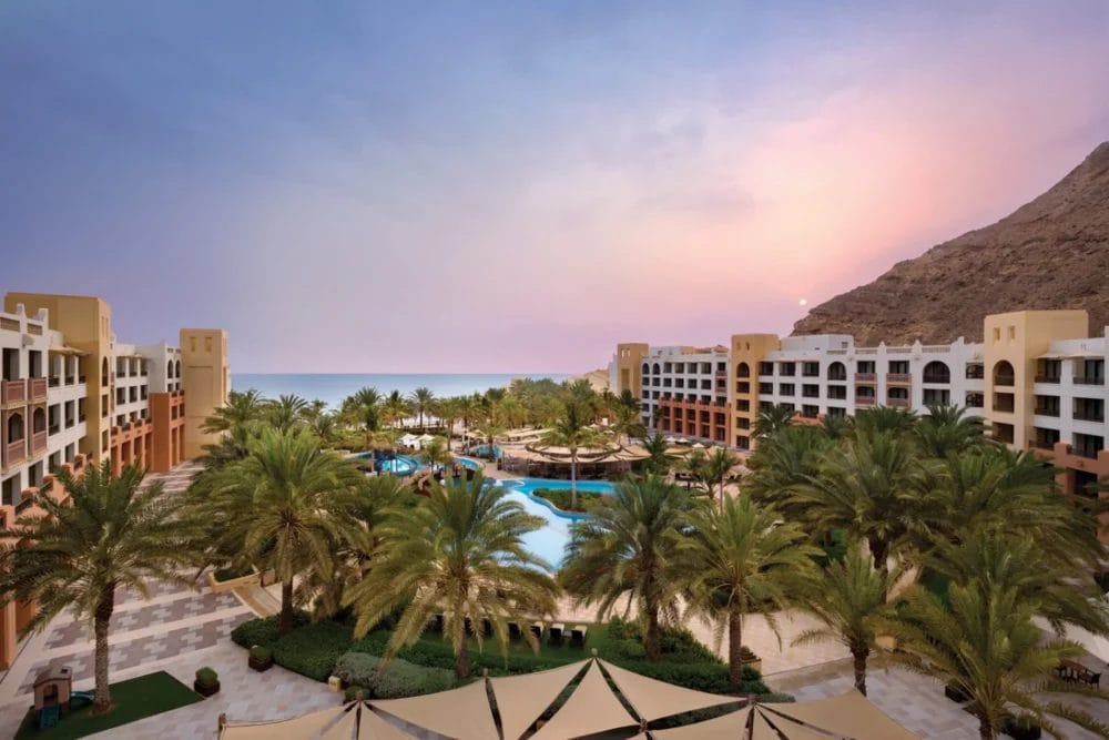 Un lussuoso resort sul mare a Barr Al Jissah con alte palme, una grande piscina ed edifici abbronzati, sullo sfondo di una montagna al tramonto e con l'oceano in lontananza: provate la tranquillità di Al Waha by Shangri-La.