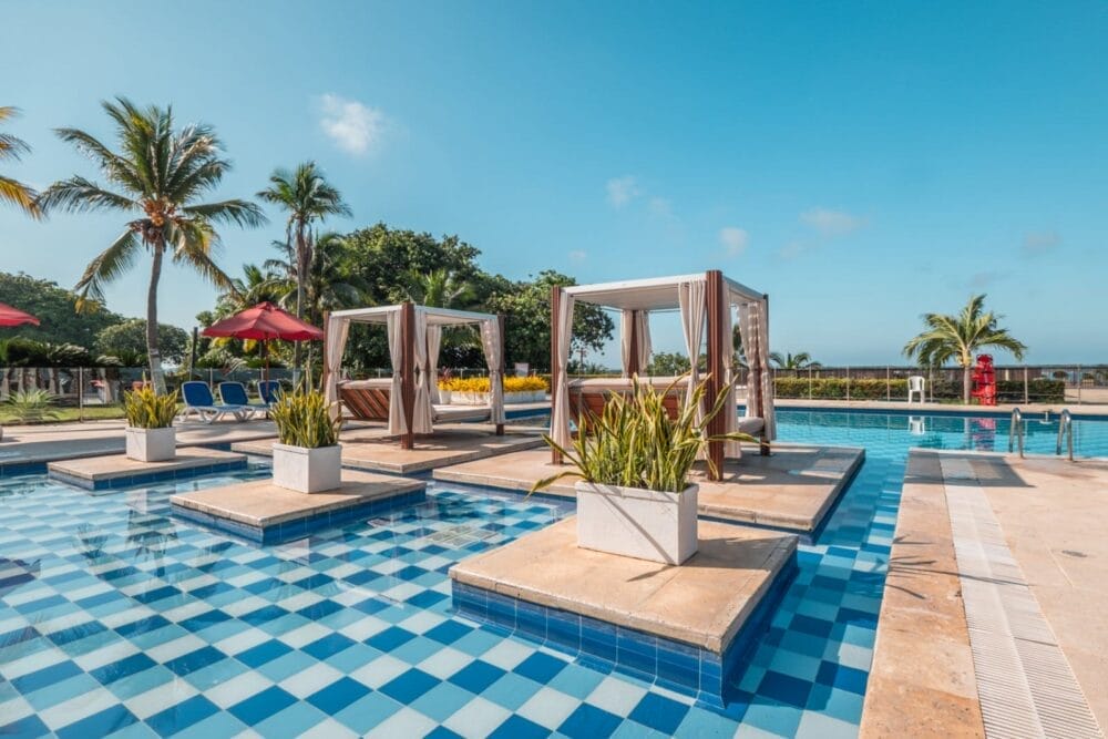 La lussuosa piscina all'aperto del Bravo Decameron Baru è caratterizzata da piastrelle blu a scacchiera, cabanas bianche su piattaforme, piante in vaso, ombrelloni rossi, palme e un cielo azzurro sullo sfondo.