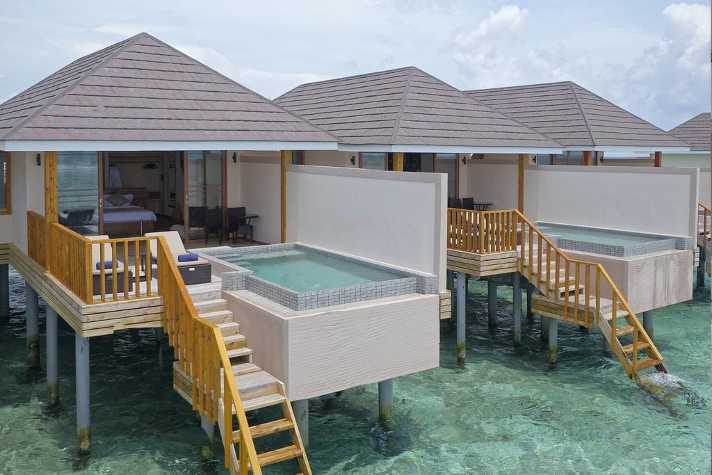 Le ville overwater con piscine private, ponti in legno e scale conducono direttamente alle acque turchesi dell'oceano in questo resort di lusso alle Maldive. Ogni villa del Brennia Kottefaru dispone di aree lounge all'aperto e tetti inclinati sotto un cielo parzialmente nuvoloso.