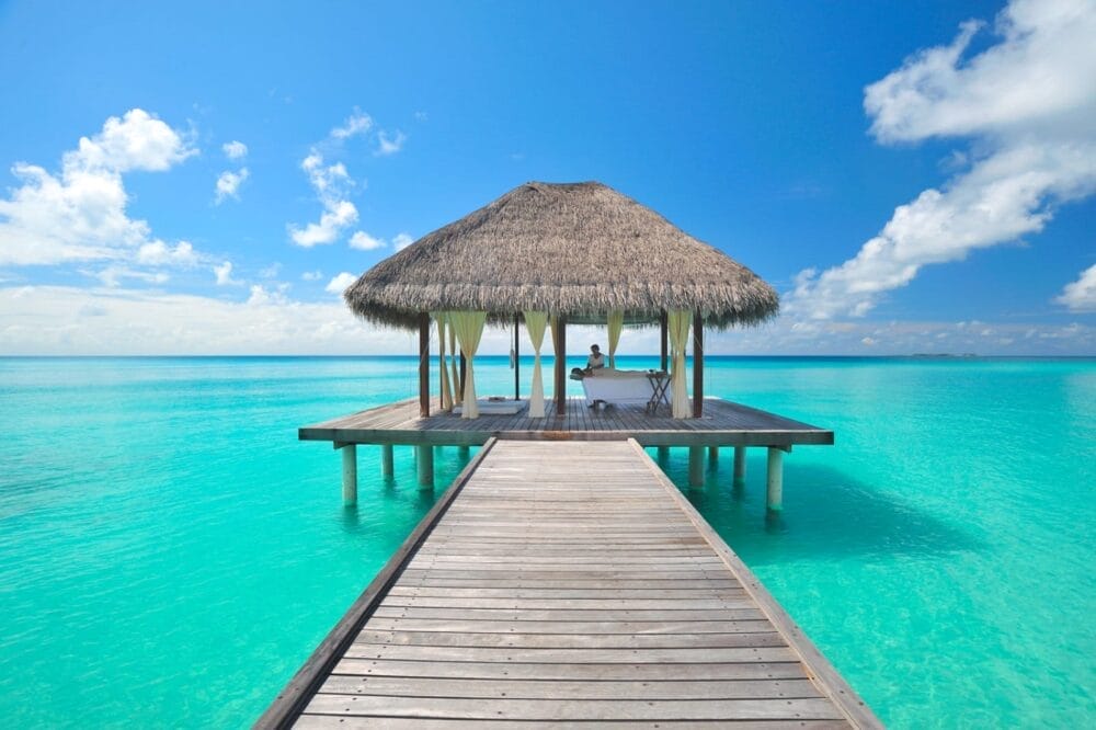 Un pontile di legno conduce a un gazebo con tetto di paglia sull'acqua limpida e turchese di Kuramathi Maldives, con una persona seduta a un tavolo, circondata da un cielo blu brillante e nuvole sparse.
