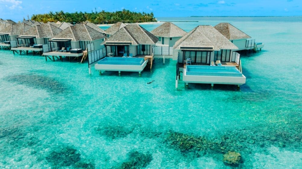 File di bungalow overwater con piscina privata si trovano sopra l'acqua limpida e turchese di un resort delle Maldive, con la barriera corallina visibile in basso e le palme dell'isola di Vakarufalhi sullo sfondo sotto un cielo azzurro.