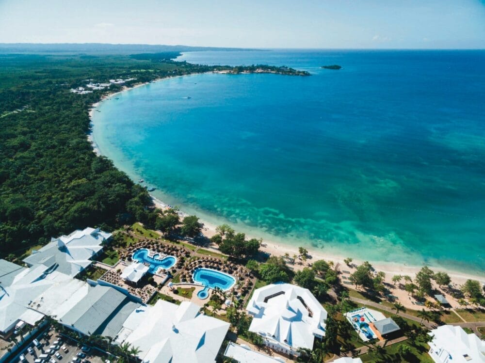 Vista aerea di una costa tropicale con acqua turchese, spiaggia di sabbia bianca, alberi verdi e un resort con piscine all'aperto, palme ed edifici dai tetti bianchi lungo la costa.
