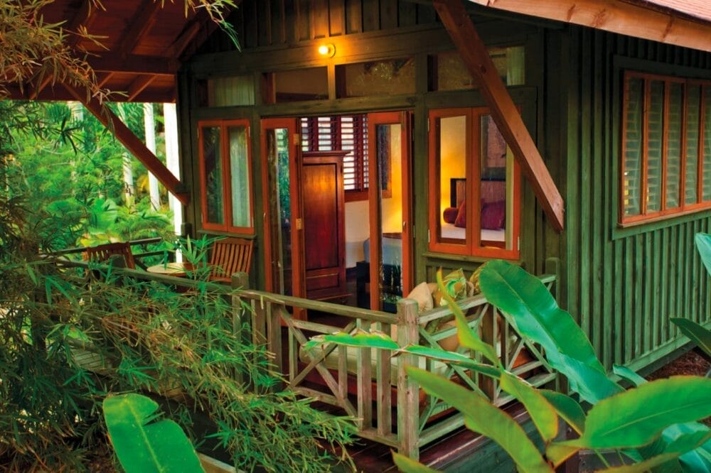 Un'accogliente cabina in legno con porte e finestre aperte, circondata da piante e alberi verdi al Sunset At The Palms. All'interno si accende una luce calda e un piccolo portico con una sedia è visibile nell'ambiente tropicale.