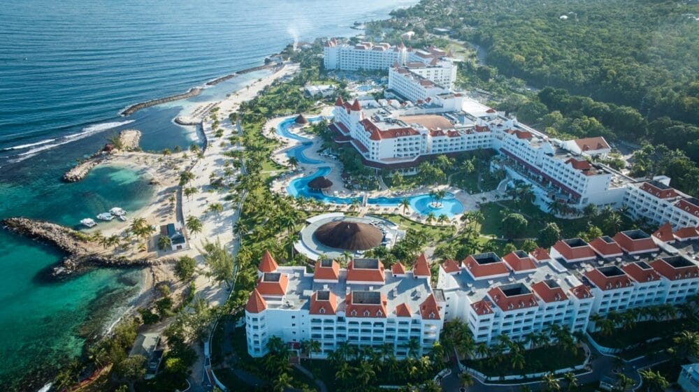Vista aerea del Bahia Principe Grand Jamaica, un hotel all-inclusive con edifici dai tetti rossi, piscine sinuose, palme e vegetazione lussureggiante accanto all'oceano blu e alle colline boscose.