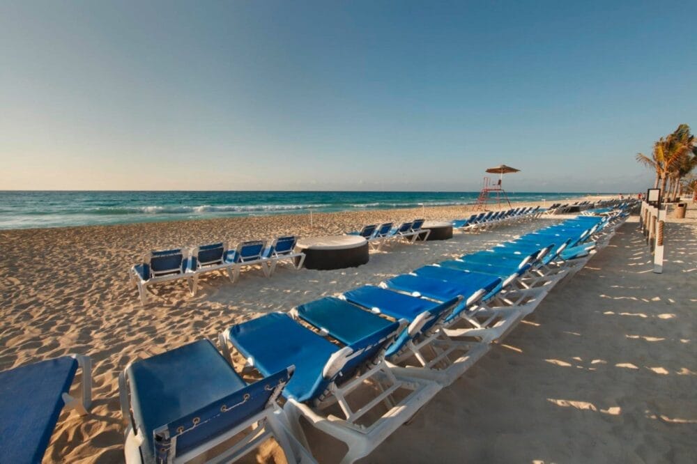 File di sedie a sdraio blu vuote sono allineate sulla spiaggia sabbiosa del resort Viva Maya, di fronte alle onde calme dell'oceano sotto un cielo limpido. Sullo sfondo si vedono una torre di salvataggio e alcune palme.