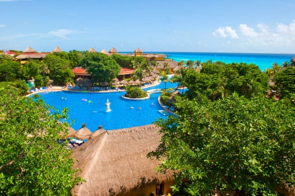 La grande piscina tropicale dell'Iberostar Waves Quetzal è circondata da alberi verdi e lussureggianti, edifici con tetti di paglia e bagnanti, con l'oceano turchese e il cielo blu sullo sfondo.