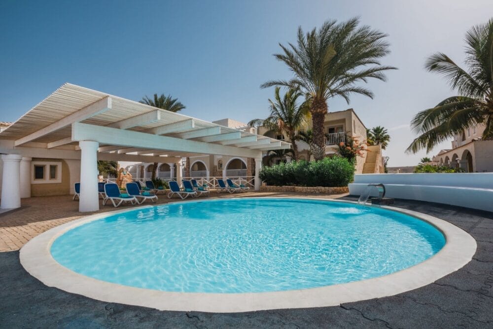 Una piscina rotonda all'aperto con acqua cristallina, circondata da sedie a sdraio sotto un pergolato bianco al Bravo Praia De Chaves. Palme ed edifici beige completano la scena rilassante in una giornata di sole.