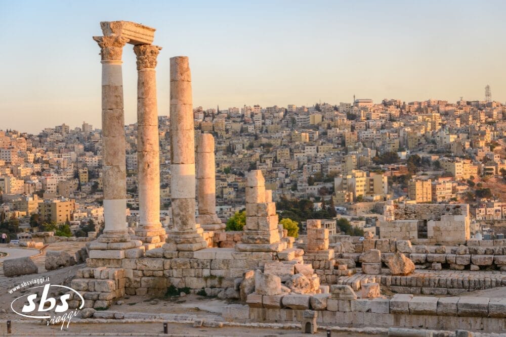 Le antiche colonne di pietra e le rovine della Cittadella di Amman sono in primo piano, mentre il paesaggio urbano si illumina al tramonto: una scena incantevole, degna di qualsiasi avventura di Magia del Deserto o di Viaggio nel Deserto.