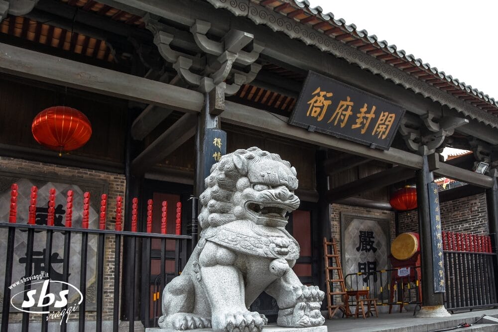 Una statua di leone in pietra si trova davanti a un edificio tradizionale cinese con lanterne rosse e insegne in legno con caratteri cinesi: una scena invitante in un Tour Cina storica, con tanto di ringhiere e pilastri neri all'ingresso.