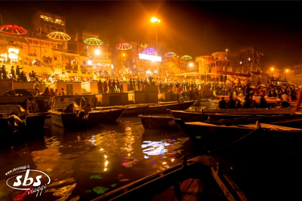 Una vivace scena notturna in un ghat sul fiume in India Splendida, con barche sull'acqua, folla radunata sui ghat e luci colorate e ombrelli che illuminano l'area durante una celebrazione festiva.