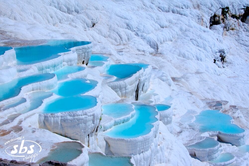 Piscine a terrazza di acqua blu brillante incastonate in formazioni bianche ricche di minerali a Pamukkale, in Turchia. Questo paesaggio unico, perfetto per una vacanza in Turchia, è modellato da sorgenti calde naturali che depositano calcio, creando splendide terrazze a gradoni.