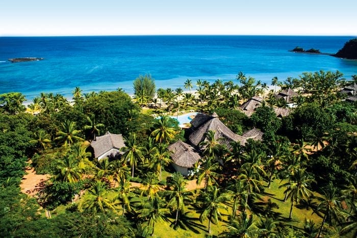 Veduta aerea del resort Amarina con palme verdi, edifici tradizionali con tetto di paglia, piscina e spiaggia sabbiosa con acqua azzurra sotto un cielo limpido, parte dell'esclusiva collezione Alpiclub.