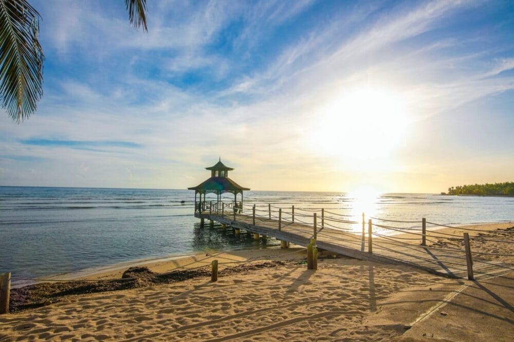 Un molo di legno con un piccolo gazebo si estende sulle calme acque dell'oceano al tramonto, con la luce dorata del sole che si riflette sulle onde vicino a una spiaggia sabbiosa. Una foglia di palma pende sopra, evocando l'atmosfera serena degli hotel di Montego Bay come il Catalonia Montego Bay.